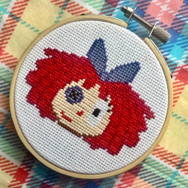Cross-stitch portrait of a red-haired character with a blue bow