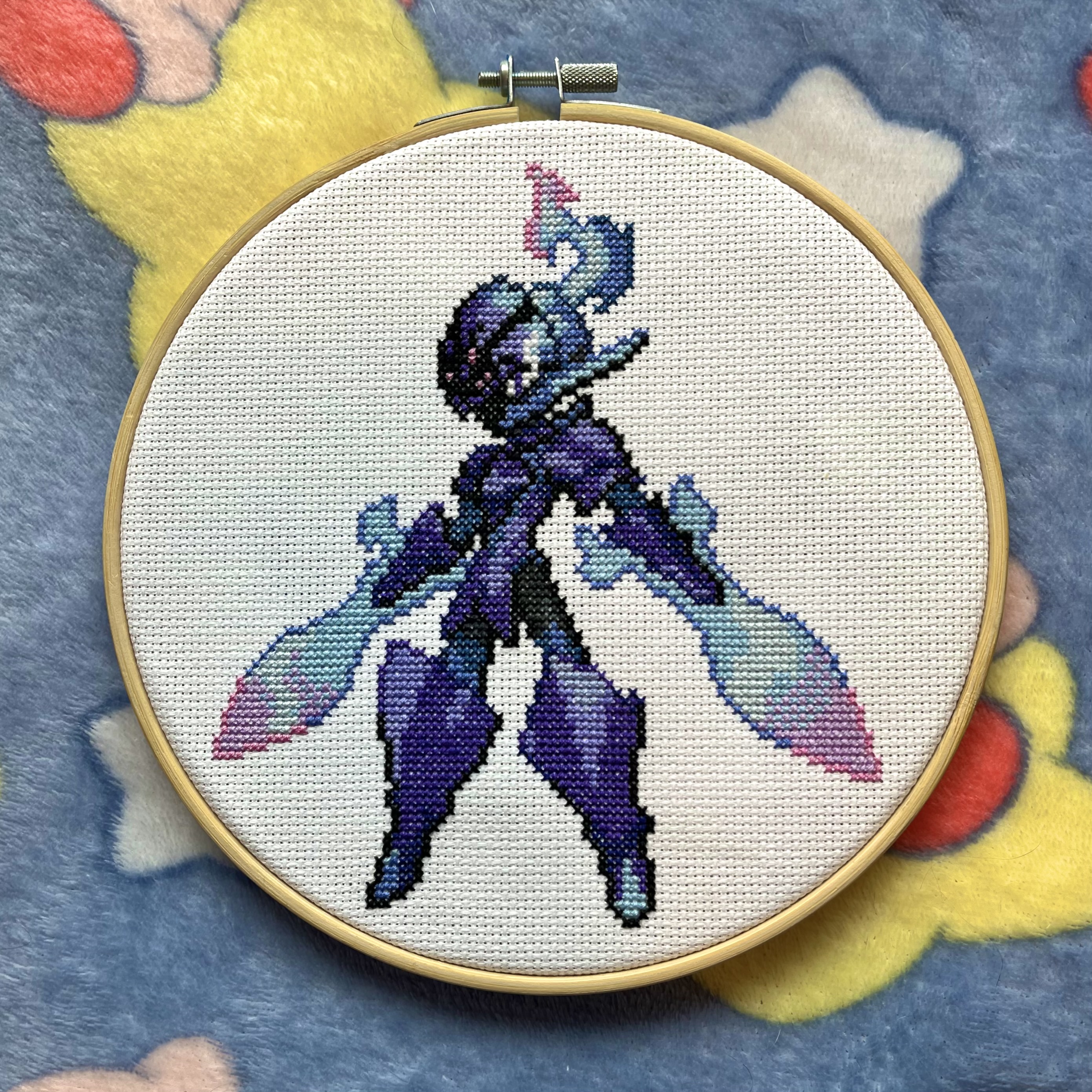 Cross-stitch of Ceruledge in a wooden hoop - the purple blade-armored Pokémon in a dynamic pose with teal blade-wings and a pink flame accent at the top, on white Aida cloth, against a star-print blue fleece background