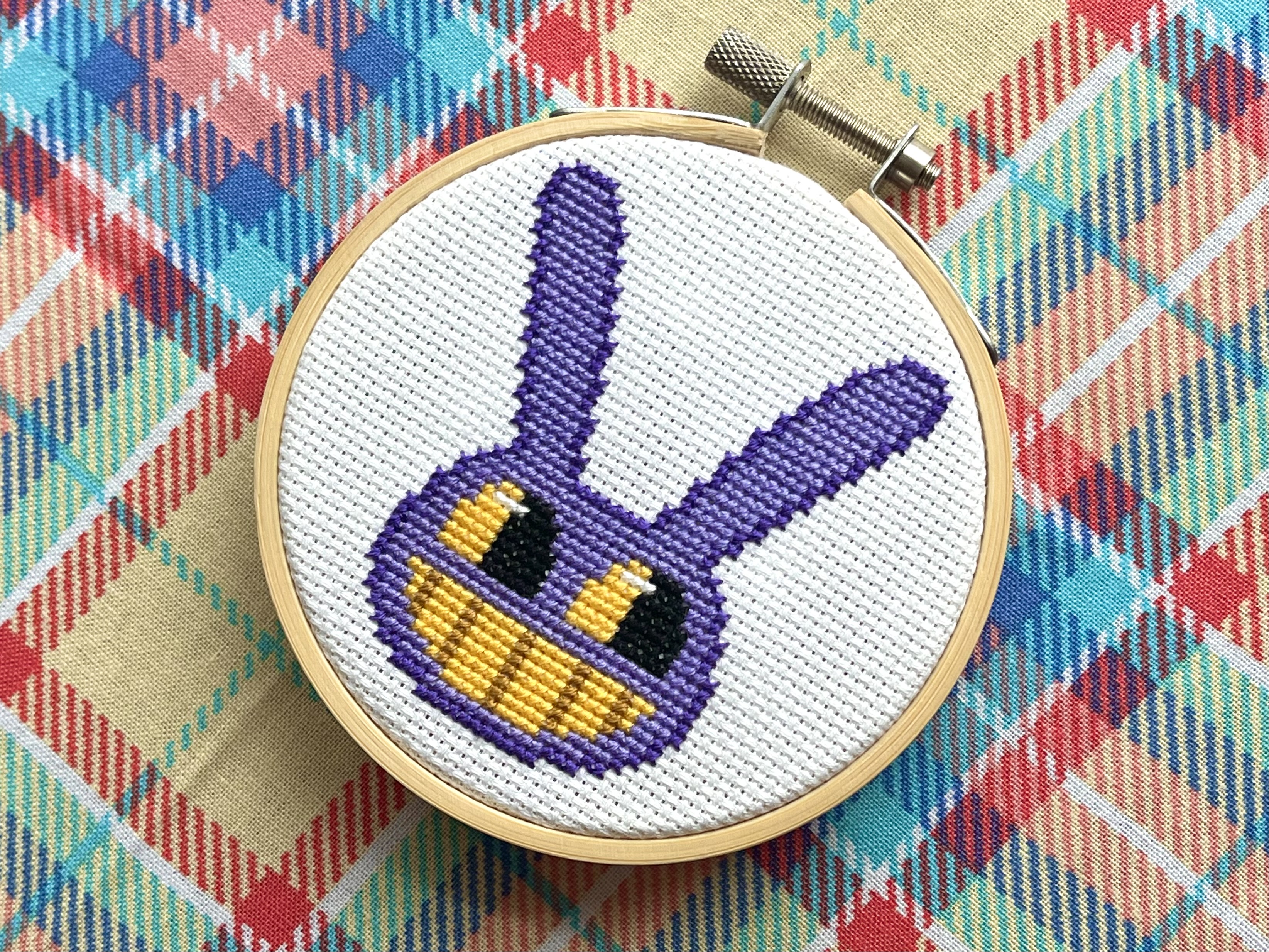 A cross-stitched portrait of Jax from The Amazing Digital Circus in a wooden embroidery hoop - a purple rabbit head with long ears, yellow eyes, and a wide toothy grin, on a colorful plaid fabric