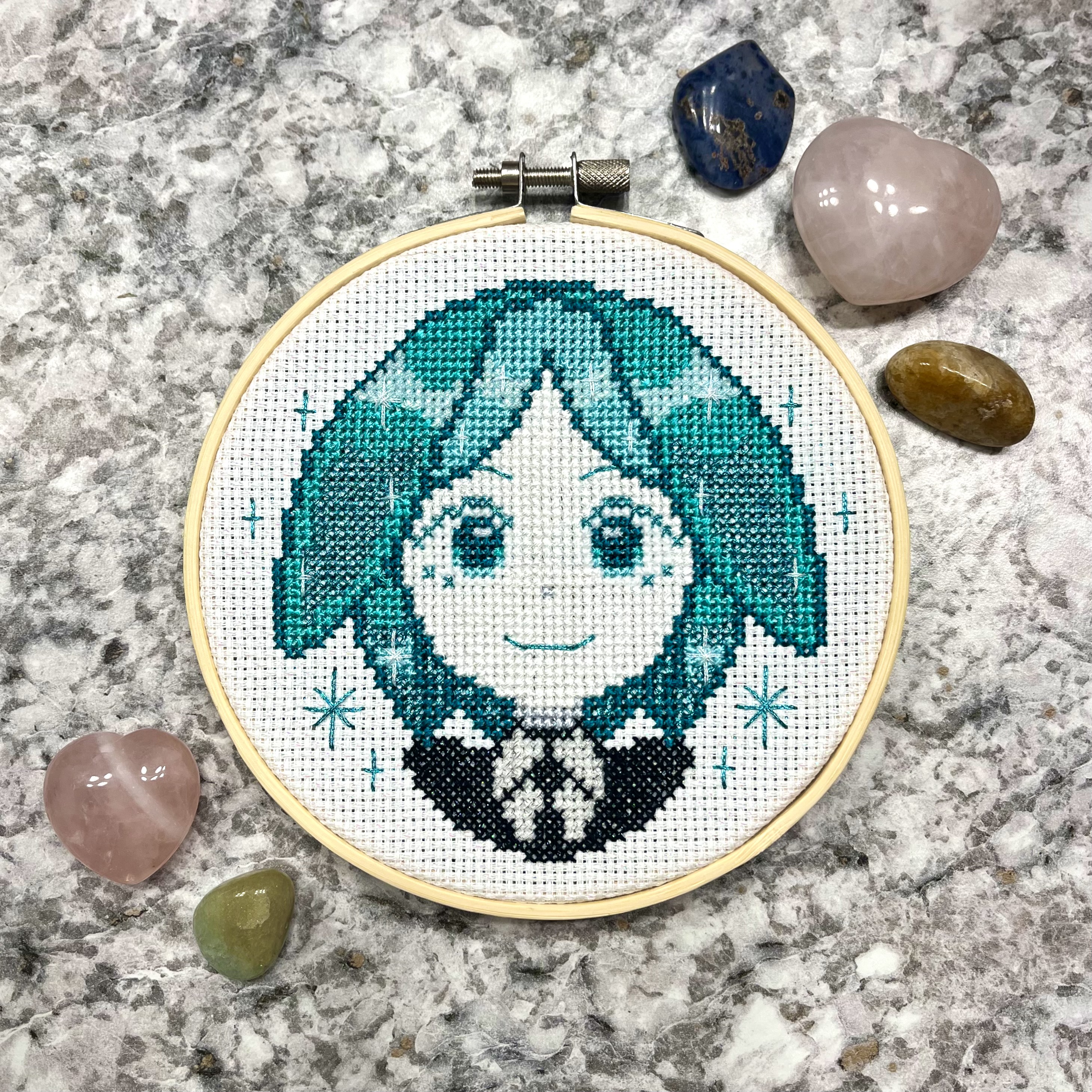 Cross-stitch of Phosphophyllite in a wooden hoop - a chibi portrait with teal hair and sparkle accents, smiling, on white Aida cloth, photographed on a marble surface with tumbled crystals arranged around the hoop