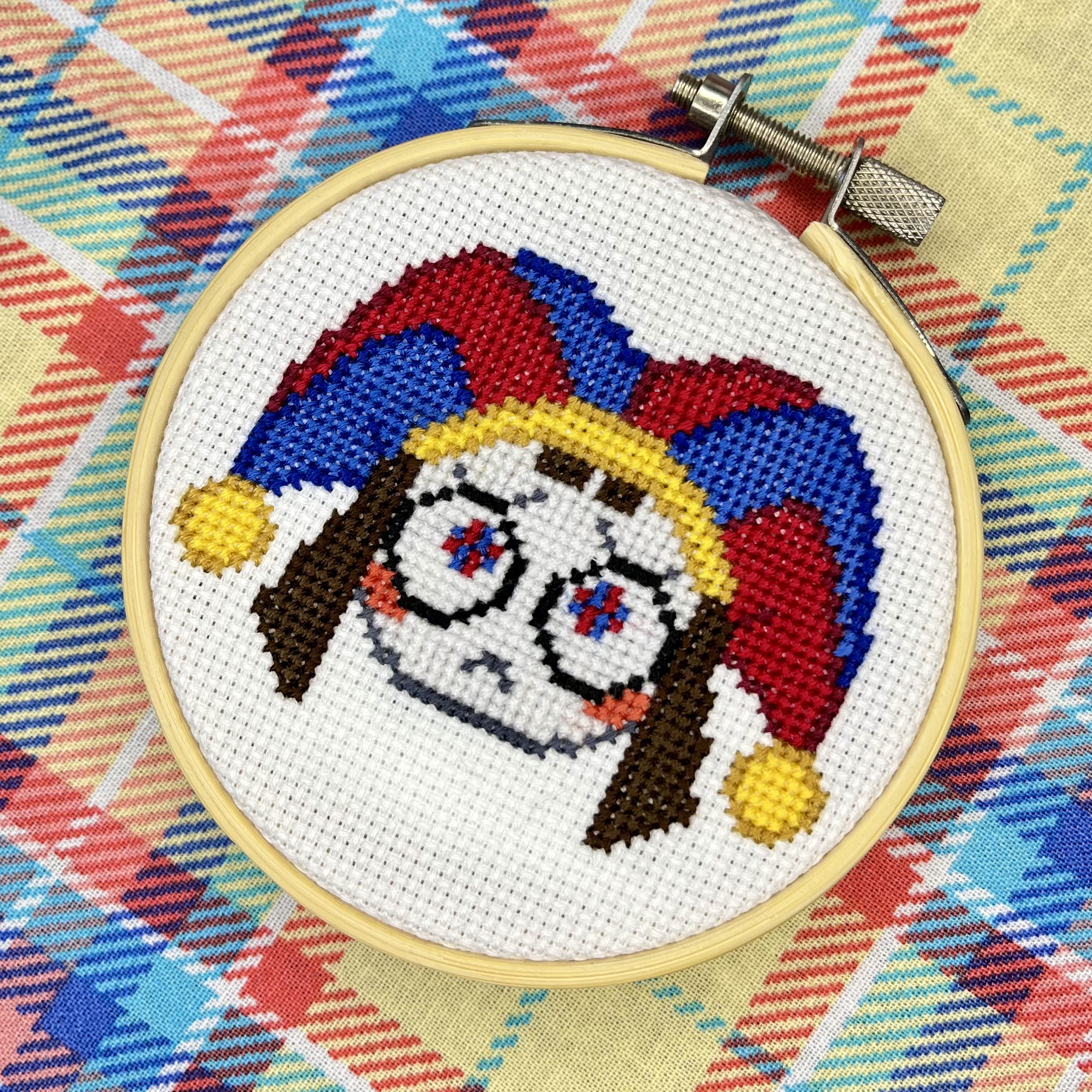 A cross-stitched portrait of Pomni from The Amazing Digital Circus in a wooden embroidery hoop - jester hat in red and blue with yellow bells, brown pigtails, and wide circular eyes with star details, on a colorful plaid fabric
