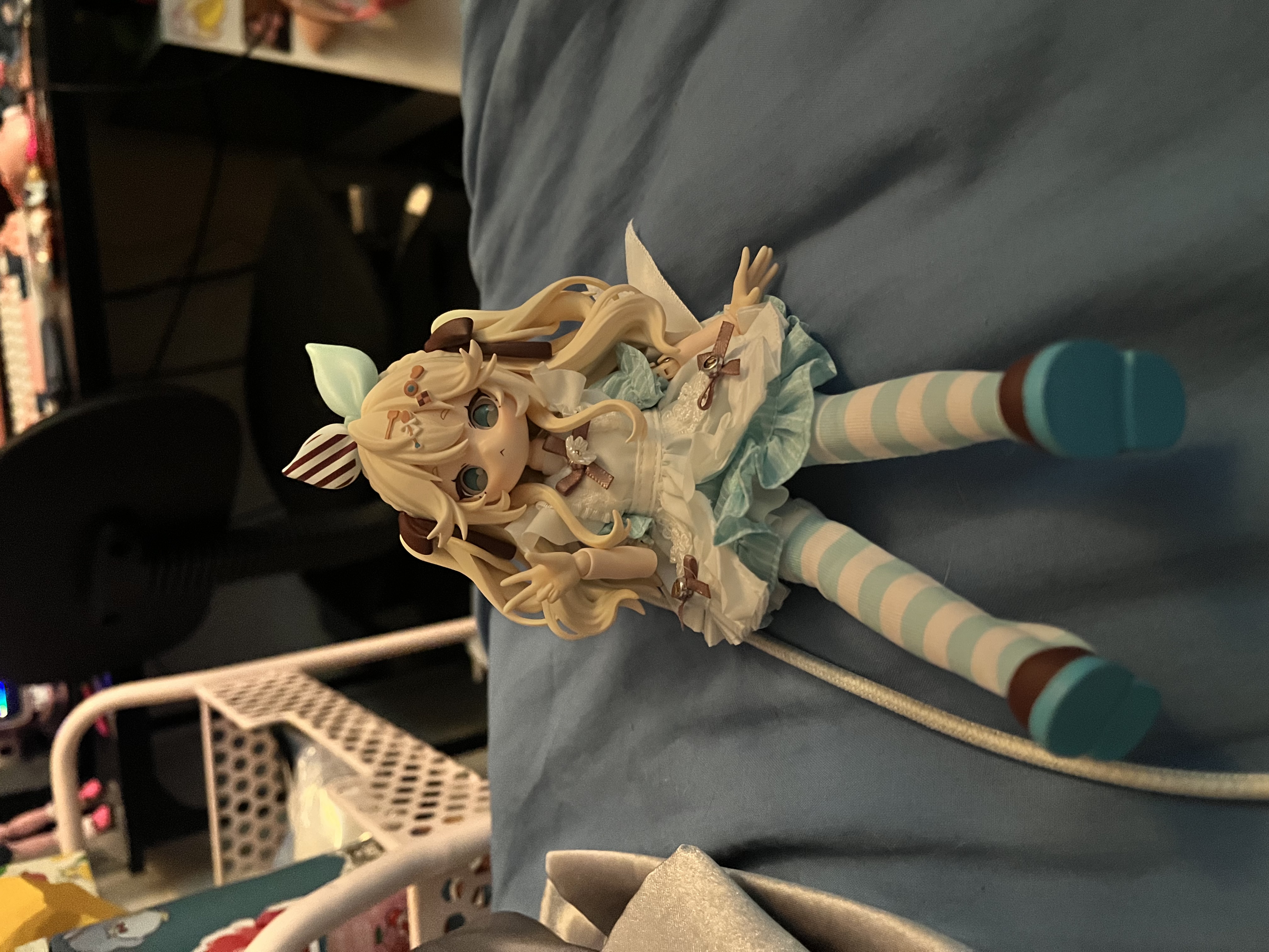 The Alice Sugary Girls doll lying on a blue fabric surface - small articulated figure with long blonde hair, teal and white striped stockings, frilly white dress, and chocolate-striped bunny-ear hair clips