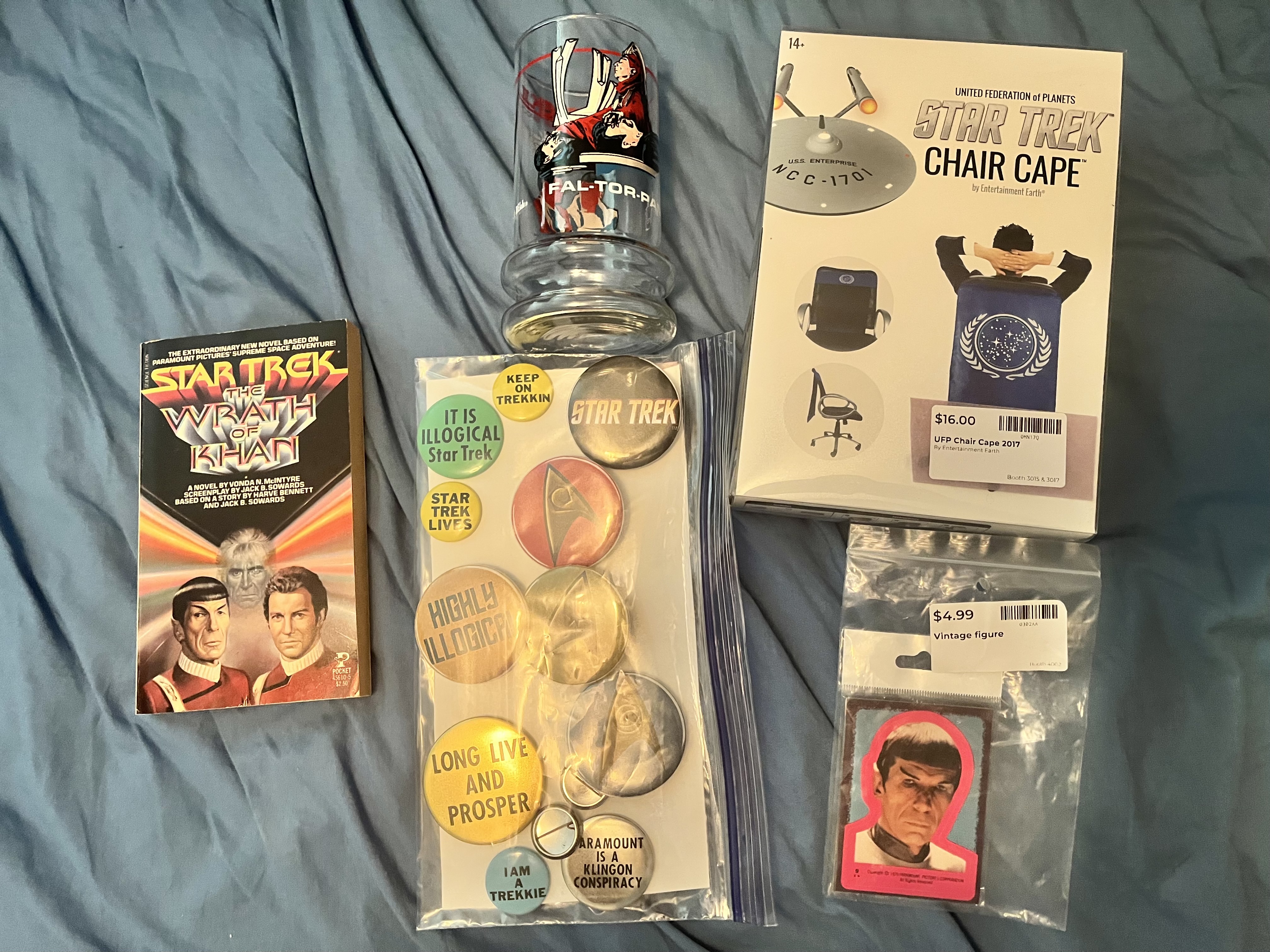 Star Trek haul spread on a blue surface: Wrath of Khan novelization, a bag of Star Trek pin buttons, the Taco Bell glass, a "Star Trek Chair Cape" box, and a vintage Spock figure in packaging
