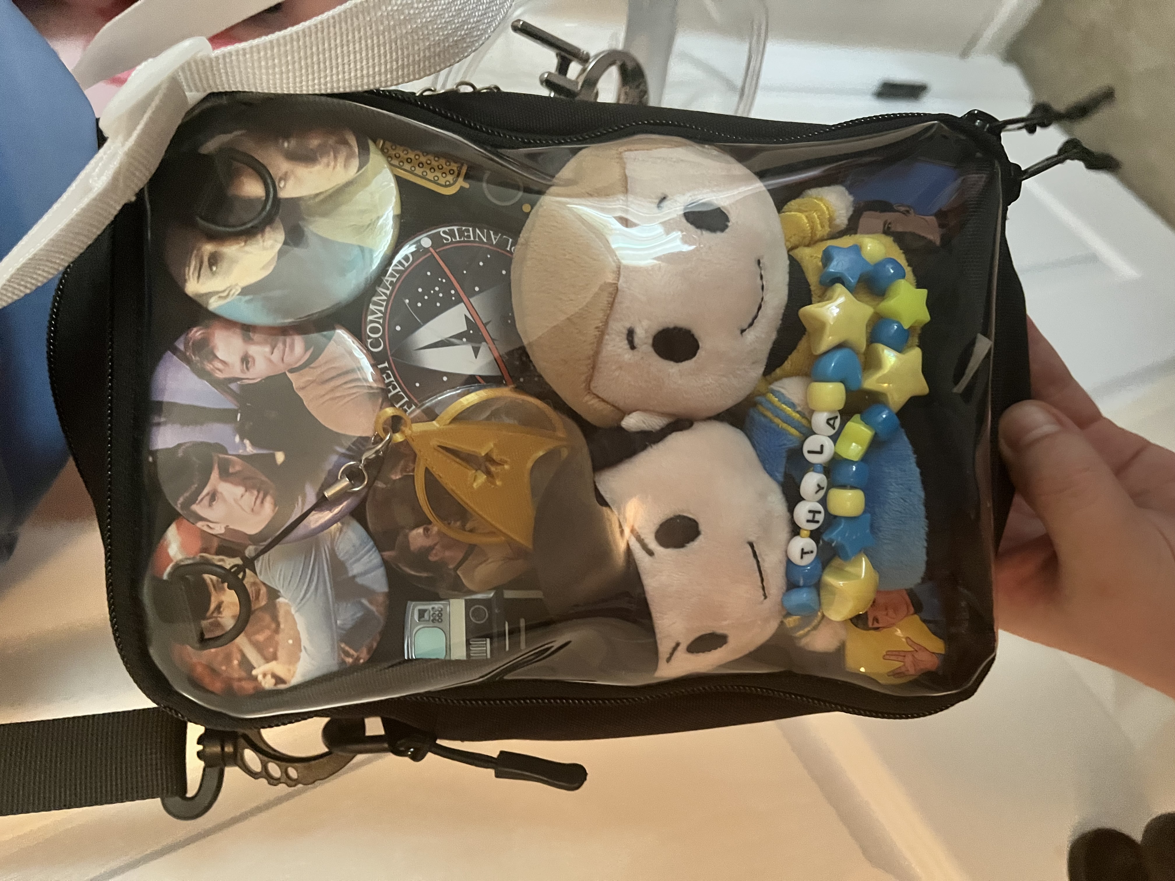 Star Trek Spirk ita bag held up - a black see-through zipper bag with TOS cast photo backing, packed with two Spock plushies, a gold Spock pin, large crew pins, and a blue-and-yellow star-bead bracelet